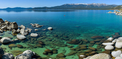 Crystal Rocky Lake - A panoramic view of colorful crystal-clear water at a rocky cove of Sand Harbor, with high peaks of Sierra Nevada at horizon, on a calm Spring evening. Lake Tahoe, CA-NV, USA.