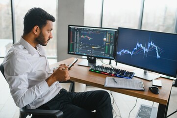 Side view of smart Indian crypto trader, investor, analyst broker, using laptop and smartphone analyzing digital cryptocurrency exchange, stock market charts, thinking of investing and funds risks.