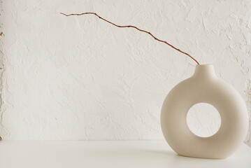 Modern beige ceramic vase with dry  branches on white table near a white textured concrete wall....