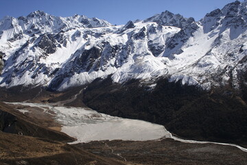 Una cadena montañosa con varios picos donde se ve la nieve entre rocas, muy escarpado y que termina en un valle con un río, con una parte helada y entre medias vegetación, en un día soleado, en Nepal