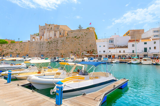 Boats line the picturesque marina port harbor at Ciutadella de Menorca, Menorca, Spain, in the Mediterranean Sea, with the historic walled fortified town and village in view on a sunny day.	