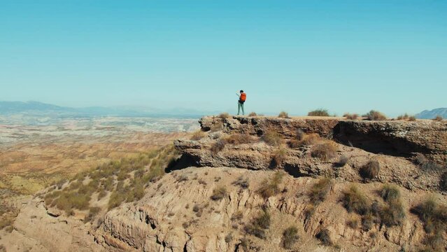 Male adventurer, surrounded by stunning scenic beauty, stands atop a desert hill. His hike to the top was worth it for the peaceful view of mountains in the distance. Aerial shot.