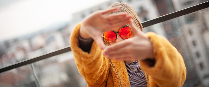 Capture Moments. Young girl in a yellow fur coat and red glasses creates a frame with hands, city in background.