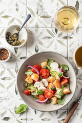 Traditional italian tomato salad panzanella with mozzarella, capers, red onion, croutons, cucumbers and basil with fork and wine glass. Summer salad on printed tile background top view