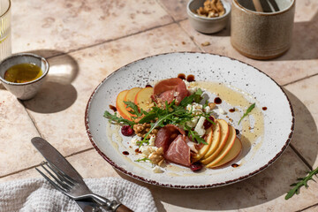Salad of arugula and prosciutto or ham, sliced apple, cheese, walnuts on rustic plate in composition with cutlery on pink tile background in hursh light