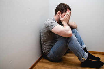 depressed man sitting in a corner of a room and covering his face