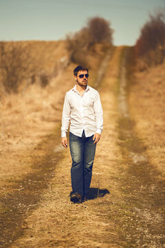 Caucasian Man Walking Down A Path In The Country Wearing Jeans, A White Button Up Shirt And Sunglasses. Looking Away From Camera