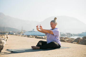 Naklejka na ściany i meble Senior woman with dreadlocks in stretching position by the sea at morning. Elderly woman doing yoga near beach.