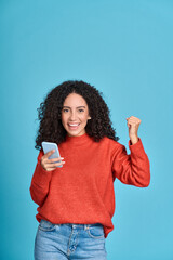 Young happy latin woman winner holding mobile cell phone isolated on blue background. Excited...