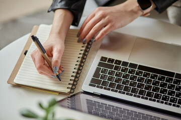 Close up woman hand using laptop and making notes in notebook sitting at workplace at home, remote work