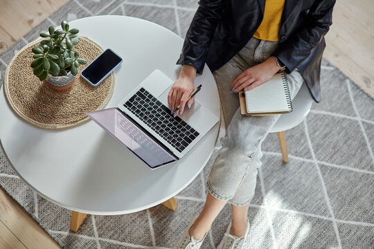 Young woman using laptop and making notes in notebook sitting at workplace at home, remote work top view