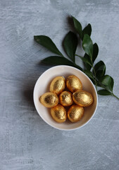 Chocolate Easter Egg Wrapped in Gold Foil. White spring flowers and Ester sweets top view photo. Bright spring still life. 
