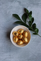 Obraz premium Chocolate Easter Egg Wrapped in Gold Foil. White spring flowers and Ester sweets top view photo. Bright spring still life. 