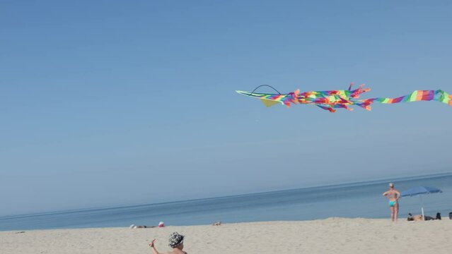 little kid boy running on beach trying to fly colorful kite in sky. bright striped kite flying in blue sky kid holding string run on white sand seaside near sea ocean edge. children summer activity 