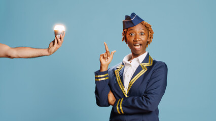 Aircrew member switching on lightbulb with new idea, feeling thoughtful over blue backdrop. Young stewardess in flying uniform lighting bulb on camera, brainstorming ideas in studio.