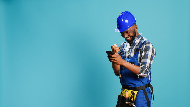 Happy construction worker using smartphone on camera, feeling confident about success. Young smiling carpenter with hardhat browsing online page and texting on mobile phone app.