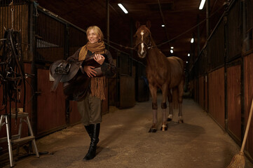 Female horse rider walking with harness in stable