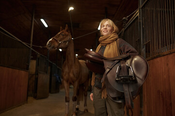 Woman rider standing and holding saddle in hand over her horse in stable