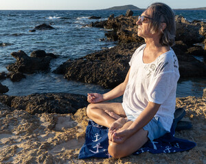 A gray-haired man with glasses practices yoga on the sea's rocky shore against the backdrop of the sea, Greece, Crete