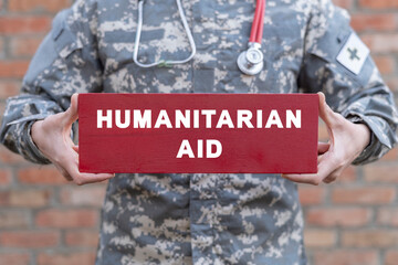 Soldier with stethoscope holding red banner with inscription: HUMANITARIAN AID. Help refugees, volunteering and rescue during the war. Humanitarian aid army concept. Humanitarian aid day.