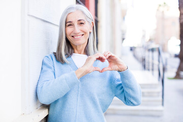 senior retired pretty white hair woman smiling and feeling happy, cute, romantic and in love, making heart shape with both hands