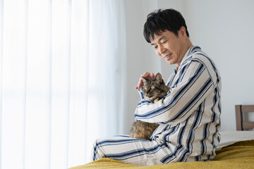 ベッドに座って猫を抱くパジャマ姿の50代男性