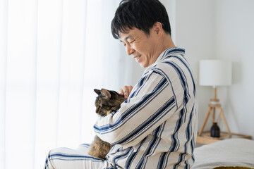 ベッドに座って猫を抱くパジャマ姿の50代男性