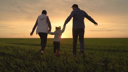 Mom, child, dad, scamper across field, happy kid holds hands of his parents, a child jumps on the green grass. Family walk in the park in spring at sunset, healthy childhood. Family weekend outdoors