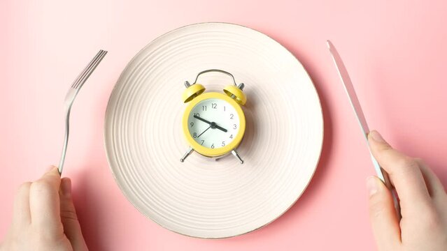 Female hands holding a fork and and knife next to an empty plate on pink background close-up top view.