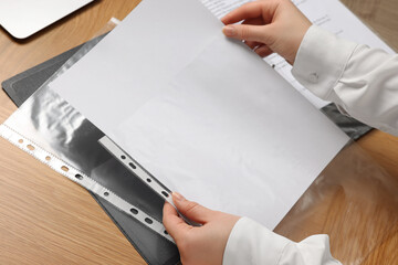 Woman putting paper sheet into punched pocket at wooden table, closeup