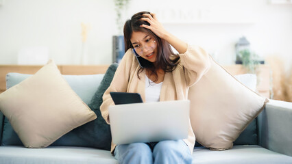 Young asian woman using computer laptop and mobile smartphone while seated on couch at home