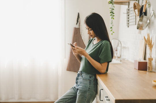 Happy young asian woman relaxing at home she is standing at counter kitchen and using mobile smartphone