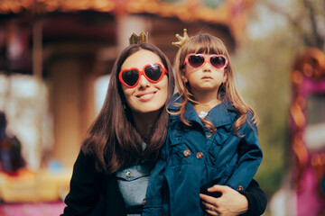 Mother and Daughter Wearing Matching Heart Shape Sunglasses and Crowns. Cute adorable mom and girl having fun in an amusement park
