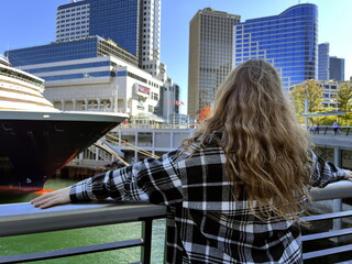 Fototapeta na wymiar A young girl looks at sea on a huge cruise liner that is sailing off the coast of Canada Canada Place Center Vancouver and skyscrapers are also visible she is in a black plaid shirt her hair is loose