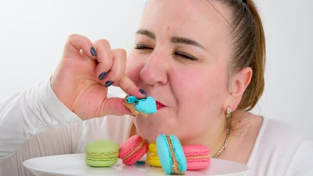 multi-colored macarons on a plate a fat woman joyfully eats them quickly licking enjoys a dessert white on a white background white clothes red lips hands diet calories overeating cand