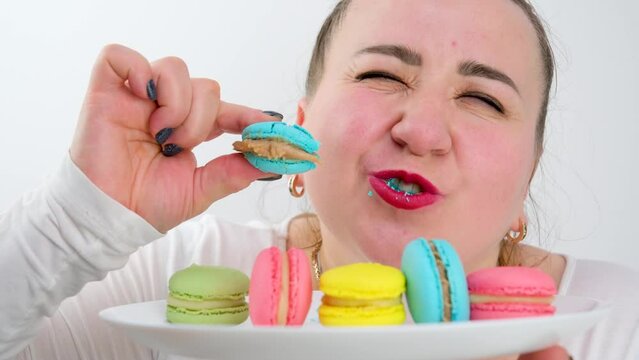 multi-colored macarons on a plate a fat woman joyfully eats them quickly licking enjoys a dessert white on a white background white clothes red lips hands diet calories overeating cand