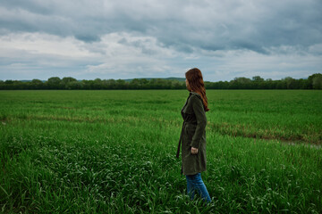 Naklejka na ściany i meble a woman with beautiful, long, red hair stands with her back to the camera in a green field in rainy, spring weather in a long raincoat