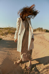 a stylish woman in a light jacket poses on the sand, turning away from the camera and the wind blows her red hair strongly