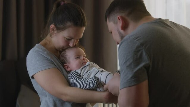 Portrait view of caring mother holding and kissing baby daughter or son in her arms. Dad plays with legs of infant in bodysuit at nursery. Concept of childhood, parenthood, family and love. Zoom in.