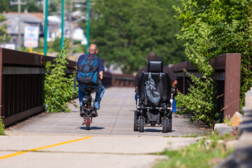 A man on a bicycle and a man in a motorized wheelchair riding down a pedestrian bridge on a sunny...