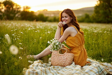 happy redhead woman sitting in a field of daisies on a plaid during sunset and smiling at the...
