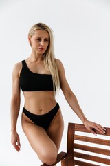 Girl in swimsuit on white wall. Beautiful young tanned slender blonde with long hair posing in studio on white background in black two-piece swimsuit. Pumped-up fitness model.