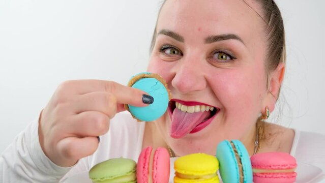 multi-colored macarons on a plate a fat woman joyfully eats them quickly licking enjoys a dessert white on a white background white clothes red lips hands diet calories overeating cand
