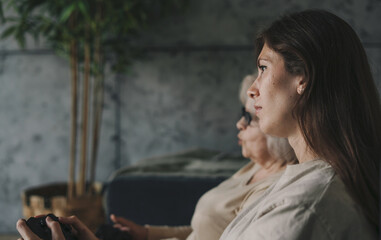 Fototapeta na wymiar Side view of a woman with her mother playing games on TV, sitting on cozy sofa. Healthy activity. Happy cheerful family. Happy lifestyle. Family home leisure.