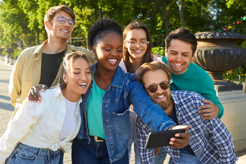 Cheerful positive young multiracial friends guys and girls making group selfie photo in summer city park looking at smartphone camera with smiles. Friendship, having fun, hanging out together concept.