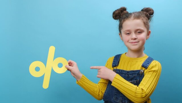 Pleasant happy little girl kid holding yellow percent mark, expressing positive emotions, presenting discounts, posing isolated over blue color background wall in studio with copy space. Sale concept