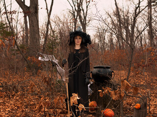 Beautiful woman in witches hat and costume holding broom near the cauldron in autumn forest. Halloween concept. Selective focus