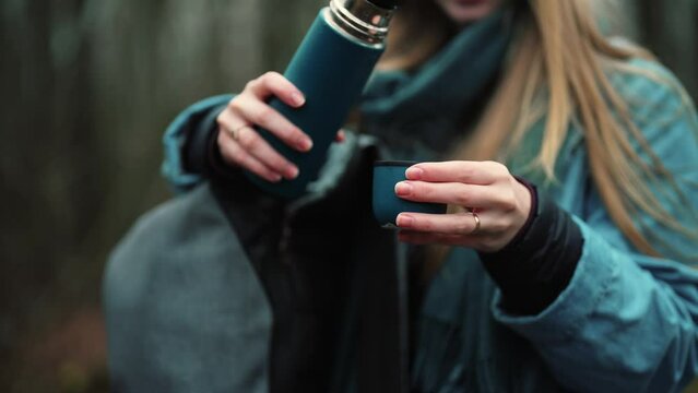 Girl hands pouring hot tea for warming from thermos in mug. Young woman brings cup to mouth and drinks it. Enjoying beautiful view of nature on forest. Autumn walking in woodland.