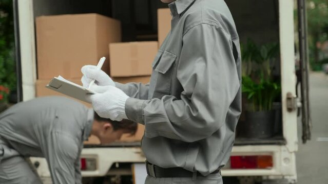 Cropped shot of moving service worker counting boxes and making notes on clipboard while his colleague unloading truck