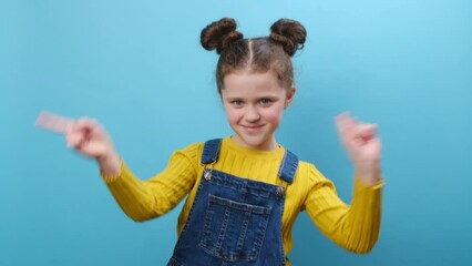 Cheerful cute funny little kid girl dancing clenching fists showing victory sign waving hands, posing isolated over blue color background wall in studio. Childhood lifestyle and people emotion concept